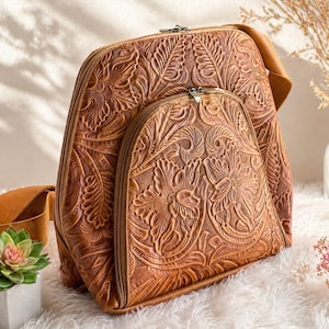 May include: A brown leather backpack with a floral embossed design. The backpack has a large main compartment and a smaller front pocket, both with zipper closures. The bag has a top handle and adjustable shoulder straps.