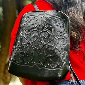 Może przedstawiać: Czarny skórzany plecak z wytłoczonym kwiatowym wzorem. Plecak ma zakrzywioną górę, złoty zamek błyskawiczny i frędzel. Torba jest noszona na ramieniu.