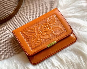 Puede incluir: Una cartera rectangular de cuero naranja con un diseño detallado de rosa en relieve en la solapa frontal. La cartera tiene costuras visibles a lo largo de los bordes y una pequeña pieza decorativa rectangular de tono dorado en el borde inferior.