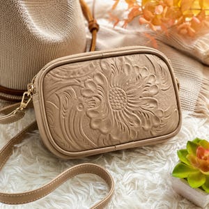 May include: A rectangular beige crossbody bag featuring an embossed floral pattern on the front panel. The bag includes a matching thin adjustable shoulder strap with visible stitching and gold-toned metal hardware attachments on the sides.