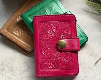 Puede incluir: Tres carteras pequeñas de cuero en rosa, verde y marrón claro, cada una con un patrón floral en relieve en la cubierta frontal. La cartera rosa está en primer plano e incluye un cierre de botón a presión color latón en una pequeña correa.