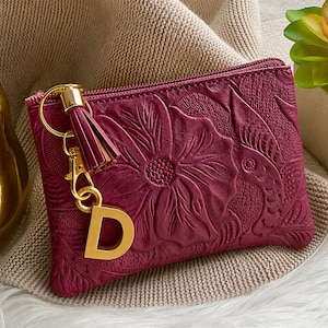 May include: A deep magenta zippered pouch featuring an embossed floral pattern on the exterior. A gold-toned metal keychain is attached to the zipper pull, consisting of a matching magenta leather tassel and a large, capital letter D charm.