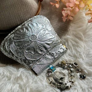 May include: A silver embossed leather wallet with a floral design. The wallet is lying on a white fluffy surface with a silver charm bracelet next to it.