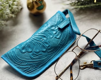 May include: A bright turquoise leather eyeglass case featuring an embossed floral pattern across the front surface. The case includes a matching turquoise strap with a gold-toned metal clasp, positioned next to a pair of thin-framed eyeglasses.