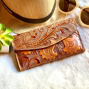 May include: A brown leather wallet with an embossed floral design. The wallet is rectangular with a flap closure. A straw hat and sunglasses are in the background. A small succulent plant is to the left.