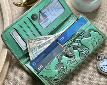 Puede incluir: Cartera de cuero verde abierta, con diseño floral en relieve, que contiene efectivo, tarjetas de crédito y un documento de identidad. La cartera está abierta, revelando múltiples compartimentos. Una figura de pájaro dorado y un pequeño reloj están en el fondo.