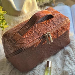 Pode incluir: Uma bolsa de cosméticos de couro marrom com um design floral em relevo. A bolsa tem uma alça superior, um fecho de zíper e puxadores dourados. A bolsa é retangular e fica em uma superfície macia e branca.