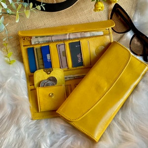 Peut inclure: Un long portefeuille en cuir jaune vif avec une fermeture à rabat. L'intérieur comprend plusieurs fentes pour cartes, un compartiment zippé et une petite poche à monnaie à bouton-pression contenant des pièces argentées. Une carte noire porte le texte well.