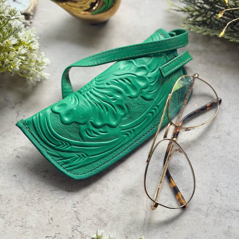 May include: A bright green leather eyeglass case featuring an embossed floral pattern and a matching strap, positioned next to a pair of gold-rimmed aviator glasses with tortoiseshell-patterned temple tips on a light gray textured surface.