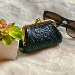 Peut inclure: Un porte-monnaie noir avec un motif floral en relief et un fermoir &agrave; cliquet en m&eacute;tal dor&eacute;. La pochette rectangulaire pr&eacute;sente des bords arrondis et une surface textur&eacute;e avec des motifs de roses et de feuilles sur le panneau avant.
