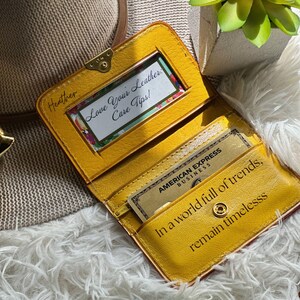 May include: A mustard yellow leather wallet featuring a clear ID window with a card reading Love Your Leather Care Tips, an American Express Business card, and the text In a world full of trends, remain timeless embossed on the interior.