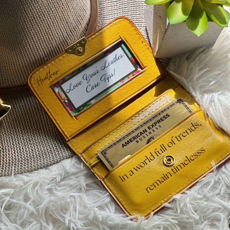 May include: A mustard yellow leather wallet featuring a clear ID window with a card reading Love Your Leather Care Tips, an American Express Business card, and the text In a world full of trends, remain timeless embossed on the interior.