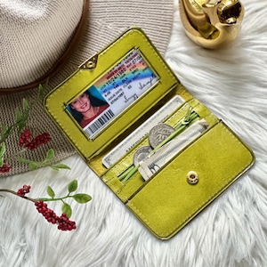 May include: A lime green bi-fold wallet featuring a clear ID window containing a sample identification card, a zippered coin compartment, and a card slot. The wallet is constructed with a metallic finish and a gold-toned snap button closure.