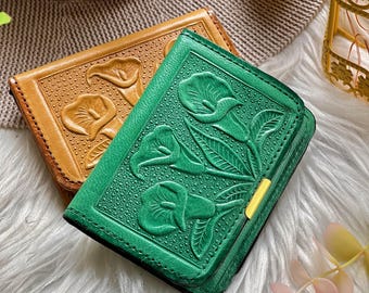 Puede incluir: Dos carteras de cuero, una verde y otra marrón. Ambas carteras presentan un diseño floral en relieve detallado. La cartera verde tiene un cierre dorado. Las carteras están sobre una superficie blanca y esponjosa.