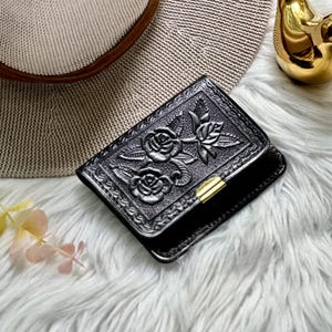 May include: A black rectangular wallet featuring an embossed floral pattern of roses and leaves on the front cover. The wallet is secured with a small, polished gold-tone metal clasp centered on the edge. The surface has a textured finish.