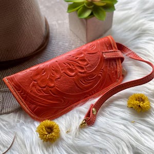 May include: A bright orange rectangular wristlet featuring an embossed floral pattern across the surface. The accessory includes a matching orange leather strap attached with a gold-toned metal rivet and a secure strap closure on the front side.