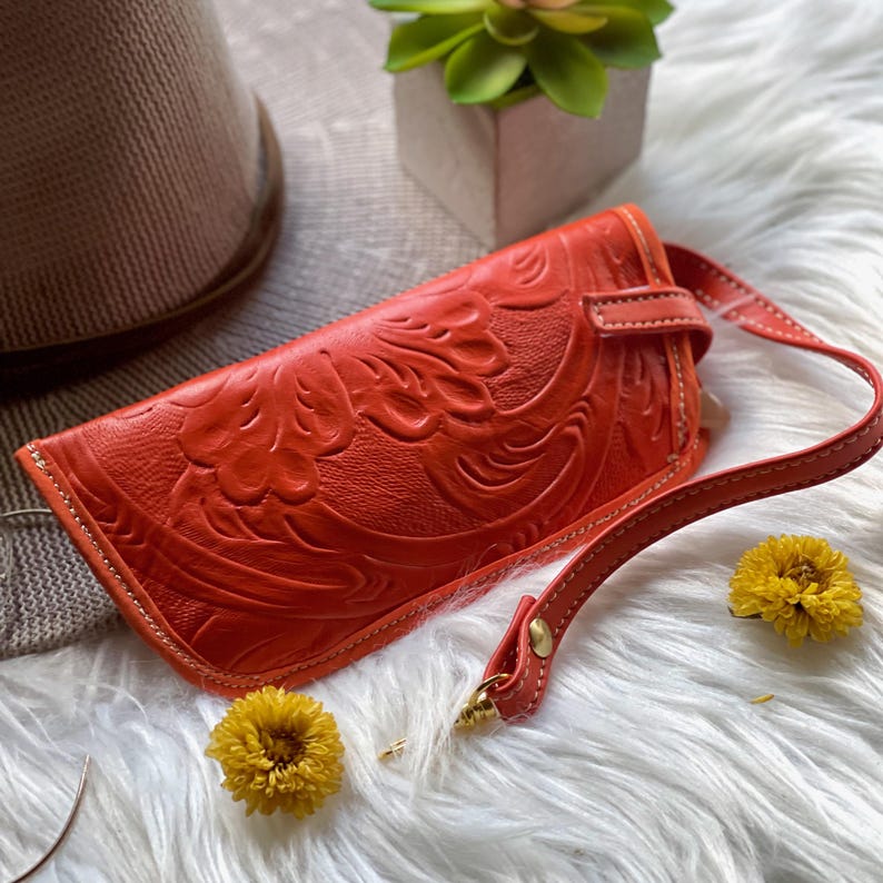 May include: A bright orange rectangular wristlet featuring an embossed floral pattern across the surface. The accessory includes a matching orange leather strap attached with a gold-toned metal rivet and a secure strap closure on the front side.