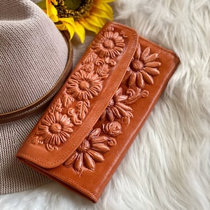 May include: A tan leather wallet with a floral design. The wallet features embossed sunflowers and leaves. The wallet is closed with a flap. A straw hat and a sunflower are in the background.
