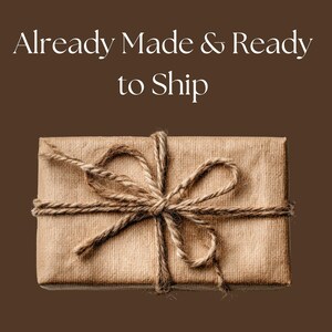 K&ouml;nnte beinhalten: Ein rechteckiges Paket, das in braunes Papier gewickelt und mit Bindfaden verschn&uuml;rt ist. Der Bindfaden ist oben zu einer Schleife geknotet. Der Text "Already Made & Ready to Ship" befindet sich oben im Bild.