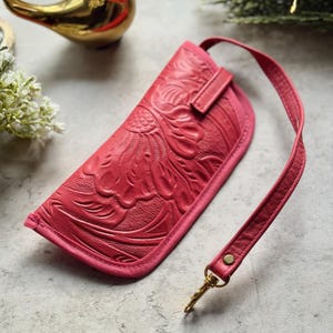 May include: A hot pink leather glasses case featuring an embossed floral pattern across the front surface. The case includes a matching pink leather wrist strap attached with a gold-toned metal swivel clasp and a small gold-toned rivet detail.