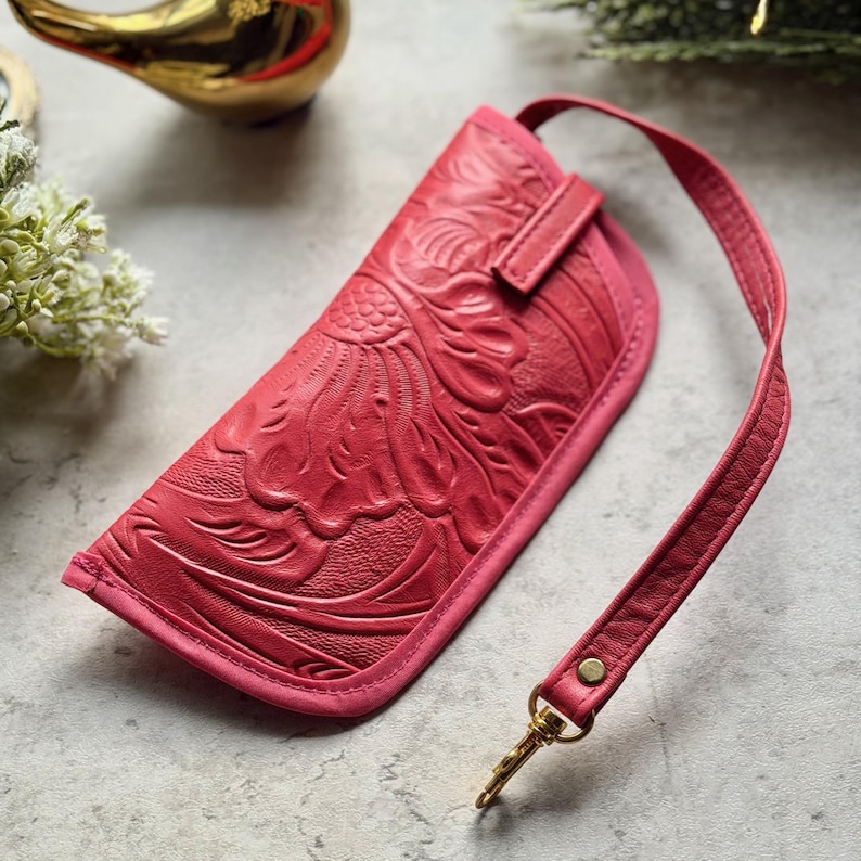 May include: A hot pink leather glasses case featuring an embossed floral pattern across the front surface. The case includes a matching pink leather wrist strap attached with a gold-toned metal swivel clasp and a small gold-toned rivet detail.