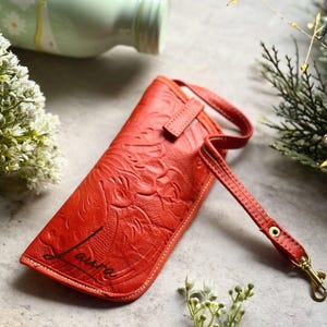 May include: A bright orange rectangular pouch with an embossed floral pattern and the name Laura printed in black script. The pouch features a matching orange wrist strap attached with a gold-toned metal swivel clasp and white stitching.