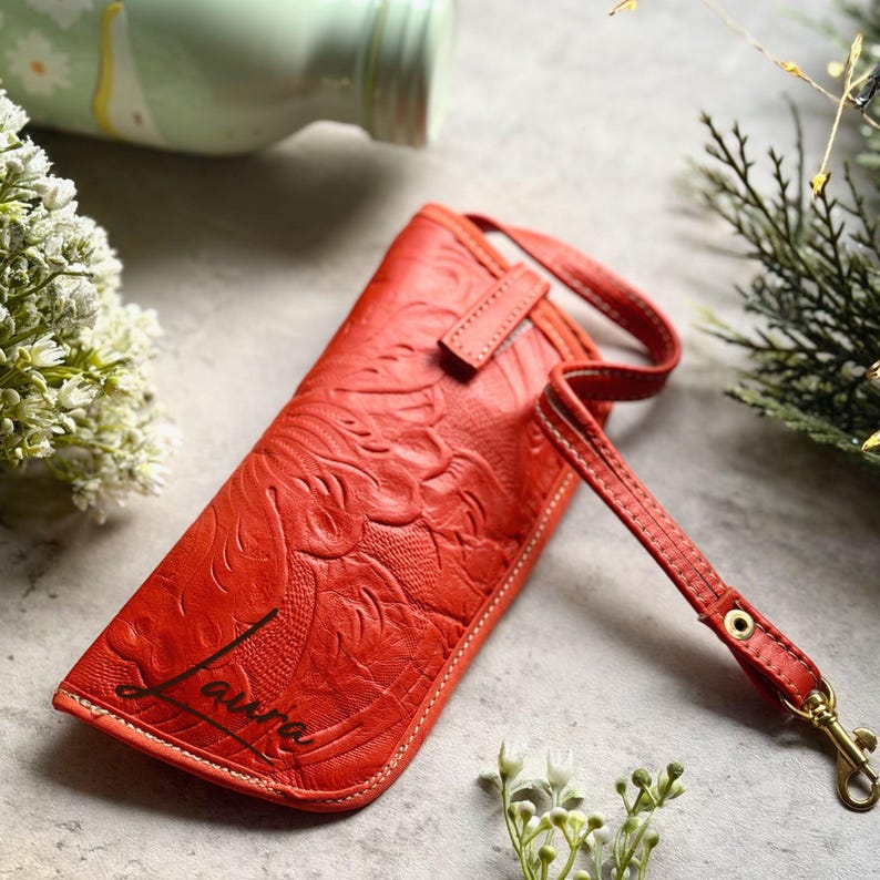 May include: A bright orange rectangular pouch with an embossed floral pattern and the name Laura printed in black script. The pouch features a matching orange wrist strap attached with a gold-toned metal swivel clasp and white stitching.