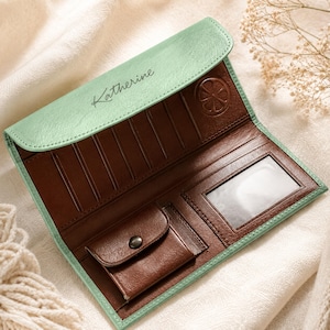 May include: A long wallet with a mint green exterior and a dark brown interior. The flap features the name Katherine in black script and a circular citrus slice emblem. The interior includes multiple card slots, a clear ID window, and a coin pocket.