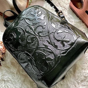 K&ouml;nnte beinhalten: Schwarzer Lederrucksack mit gepr&auml;gtem floralem Design. Der Rucksack hat einen Rei&szlig;verschluss, verstellbare Tr&auml;ger und einen oberen Griff. Das detaillierte Muster bedeckt die Vorder- und Seiten des Rucksacks.