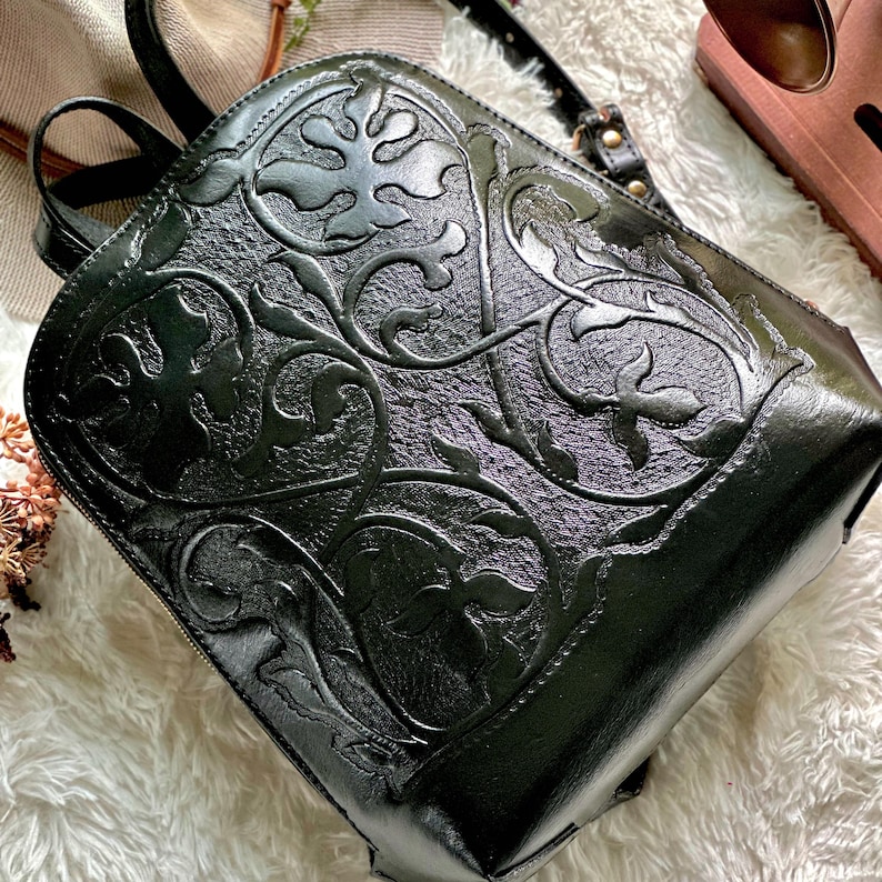 K&ouml;nnte beinhalten: Schwarzer Lederrucksack mit gepr&auml;gtem floralem Design. Der Rucksack hat einen Rei&szlig;verschluss, verstellbare Tr&auml;ger und einen oberen Griff. Das detaillierte Muster bedeckt die Vorder- und Seiten des Rucksacks.