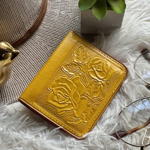 May include: A mustard yellow leather wallet featuring an embossed floral pattern of roses and leaves on the front cover. The wallet has a rectangular shape with stitched edges and a textured surface, showcasing detailed petal and foliage designs.