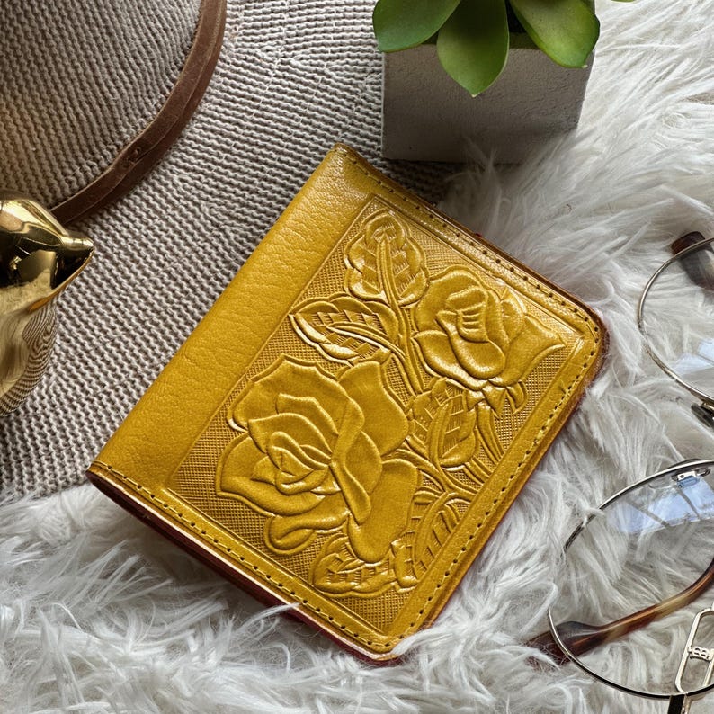 May include: A mustard yellow leather wallet featuring an embossed floral pattern of roses and leaves on the front cover. The wallet has a rectangular shape with stitched edges and a textured surface, showcasing detailed petal and foliage designs.