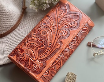 Puede incluir: Una cartera de cuero marrón con un intrincado diseño floral en relieve en la parte delantera. La cartera está cerrada y tiene una solapa.