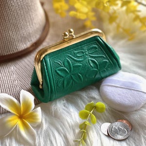 Peut inclure: Un petit porte-monnaie vert &eacute;meraude avec un motif floral en relief et un fermoir &agrave; cliquet en m&eacute;tal dor&eacute;. Le porte-monnaie pr&eacute;sente une surface textur&eacute;e et est plac&eacute; &agrave; c&ocirc;t&eacute; d'une houppette blanche, d'une fleur blanche et d'une pi&egrave;ce de monnaie.