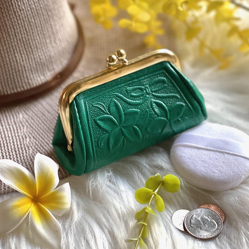 Peut inclure: Un petit porte-monnaie vert &eacute;meraude avec un motif floral en relief et un fermoir &agrave; cliquet en m&eacute;tal dor&eacute;. Le porte-monnaie pr&eacute;sente une surface textur&eacute;e et est plac&eacute; &agrave; c&ocirc;t&eacute; d'une houppette blanche, d'une fleur blanche et d'une pi&egrave;ce de monnaie.