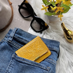 May include: A mustard yellow leather wallet featuring an embossed floral rose pattern on the front flap, tucked into the back pocket of a pair of blue denim jeans. The wallet has a rectangular shape with a small metallic accent on the bottom edge.