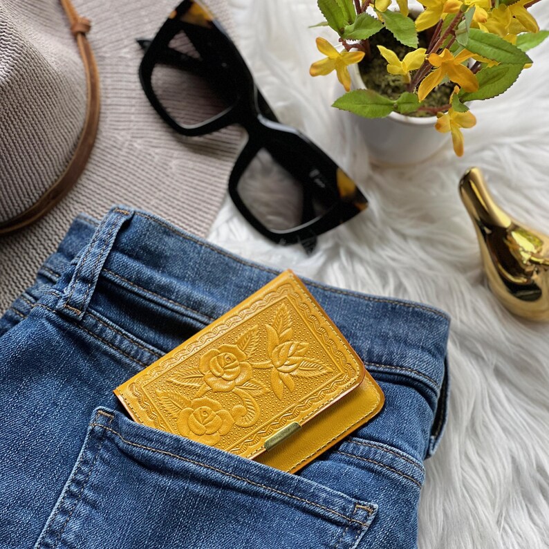 May include: A mustard yellow leather wallet featuring an embossed floral rose pattern on the front flap, tucked into the back pocket of a pair of blue denim jeans. The wallet has a rectangular shape with a small metallic accent on the bottom edge.