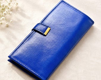 May include: A long, rectangular wallet in a vibrant royal blue color with a textured finish. The wallet features a secure strap closure across the front, accented with a small, polished gold-tone metal rectangular detail on the strap.