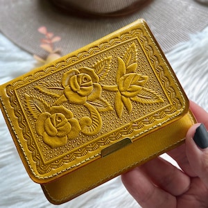 May include: A mustard yellow wallet featuring an embossed floral pattern of two roses with leaves. The rectangular wallet has a decorative scalloped border and a small metallic gold-toned rectangular accent piece centered on the bottom edge.