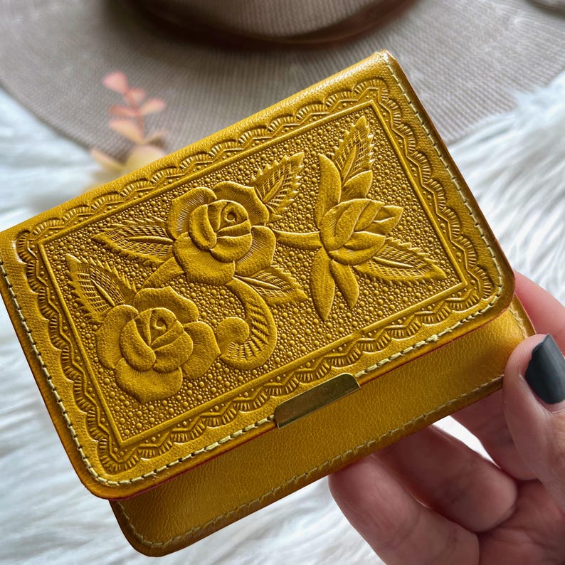 May include: A mustard yellow wallet featuring an embossed floral pattern of two roses with leaves. The rectangular wallet has a decorative scalloped border and a small metallic gold-toned rectangular accent piece centered on the bottom edge.