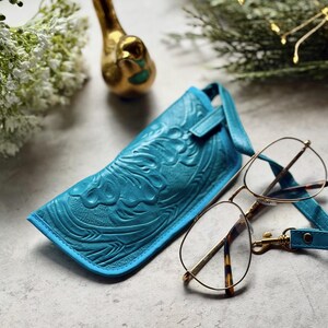 May include: A bright turquoise leather eyeglass case featuring an embossed floral pattern and a matching strap, positioned next to a pair of gold-rimmed glasses with tortoiseshell temple tips. The case has a structured shape and a smooth finish.