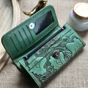 May include: Open, green leather wallet with a floral embossed design. The wallet features multiple card slots, a zippered compartment, and a snap closure. The interior includes a mirror. The wallet is open, displaying its contents.