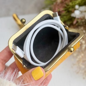 Peut inclure: Une petite pochette jaune avec un cadre m&eacute;tallique dor&eacute; &agrave; fermoir &agrave; cliquet. L'int&eacute;rieur est doubl&eacute; d'un tissu sombre et contient un c&acirc;ble de charge USB-C blanc enroul&eacute;. La pochette pr&eacute;sente une surface ext&eacute;rieure textur&eacute;e et un fermoir.