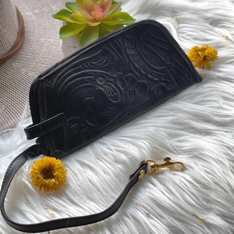 May include: A black wristlet pouch featuring an intricate embossed floral pattern across its surface. The pouch includes a matching black wrist strap attached with a gold-toned metal swivel clasp, providing a secure way to carry the accessory.