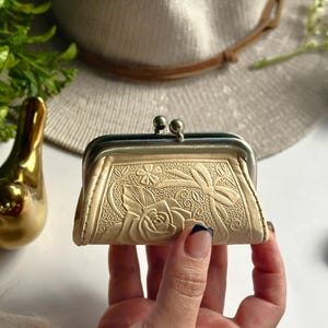Peut inclure: Un porte-monnaie de couleur cr&egrave;me avec un motif floral en relief repr&eacute;sentant une rose et un ruban. Le porte-monnaie est dot&eacute; d'un cadre en m&eacute;tal argent&eacute; avec un fermoir &agrave; cliquet sur le dessus pour s&eacute;curiser le contenu &agrave; l'int&eacute;rieur.