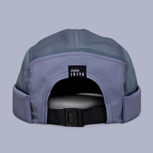Könnte beinhalten: Zweifarbiges blau-graues Cap mit gebogenem Schirm und schwarzem, verstellbarem Riemen. Die Kappe hat ein Mesh-Panel und ein kleines schwarzes Etikett mit der Aufschrift "studio IKIYO". Entworfen für eine bequeme Passform.