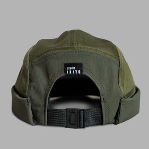 May include: Olive green cap with a curved brim and a black adjustable strap. The cap features a mesh panel and a small black tag with the text "studio IKIYO". The cap is made of a durable, textured fabric.