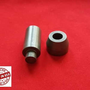 May include: Two metal tools for leather crafting. One is a round, black, metal punch with a hole in the center. The other is a silver, metal punch with a rounded top and a hole in the center.