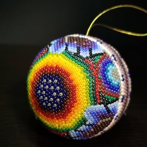 May include: A beaded ornament with a rainbow pattern. The ornament is decorated with a variety of colours, including red, orange, yellow, green, blue, and purple. A gold string is attached for hanging. The ornament is likely handmade.