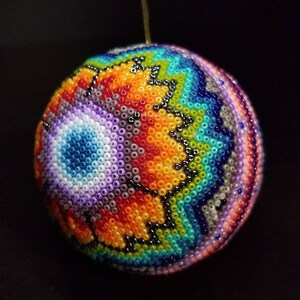 May include: A spherical ornament, intricately beaded with a rainbow of colours. The design features a geometric pattern with orange, blue, green, and purple beads. The craftsmanship suggests a handmade item, suitable for festive decorations. The diameter is approximately 7 cm.